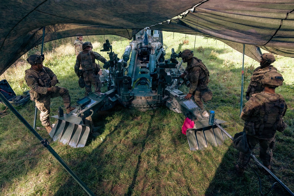 Several soldiers in camouflage uniforms are gathered around a large artillery piece, likely an M777 howitzer or similar towed howitzer, positioned under a camouflage netting canopy on a grassy field. The crew appears to be conducting maintenance, training, or preparation activities around the weapon system. The setup suggests a field exercise or operational firing position.