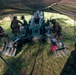 Several soldiers in camouflage uniforms are gathered around a large artillery piece, likely an M777 howitzer or similar towed howitzer, positioned under a camouflage netting canopy on a grassy field. The crew appears to be conducting maintenance, training, or preparation activities around the weapon system. The setup suggests a field exercise or operational firing position.