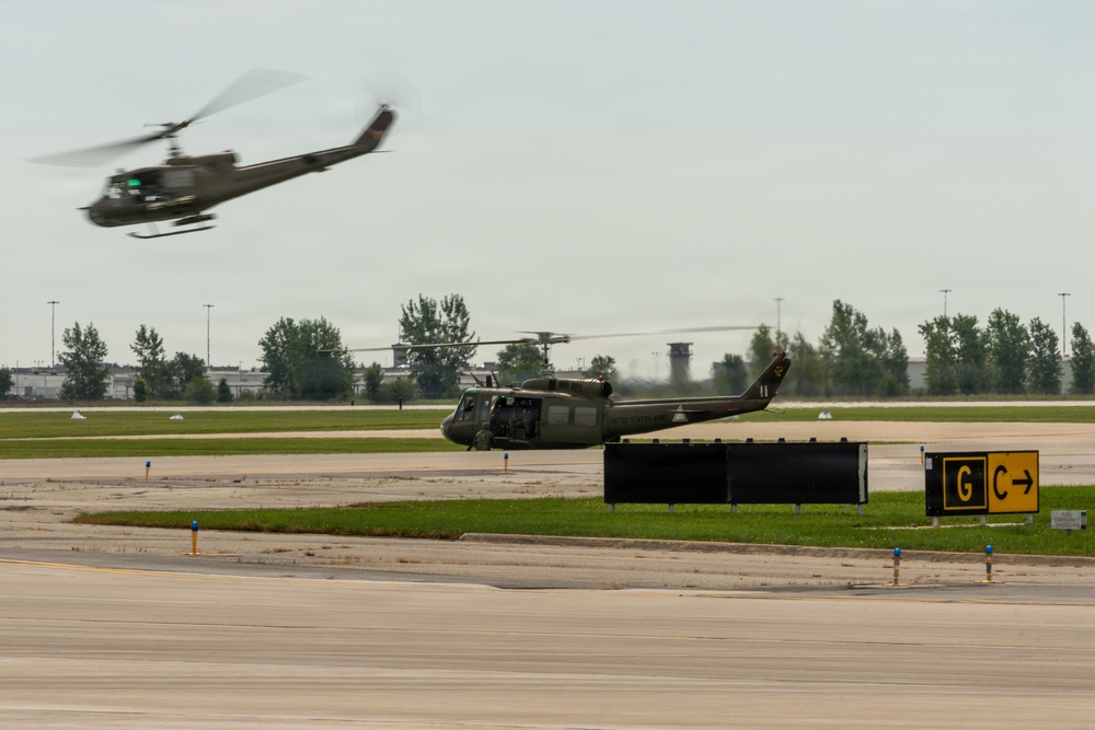 Two military helicopters, appearing to be UH-1 'Huey' variants in olive drab paint, are operating on or near an airfield tarmac. One helicopter is airborne in the upper left with motion blur indicating active flight, while the second is on or near the ground with rotor spinning. Airport taxiway signage marked 'G' and 'C' is visible in the foreground.