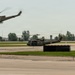 Two military helicopters, appearing to be UH-1 'Huey' variants in olive drab paint, are operating on or near an airfield tarmac. One helicopter is airborne in the upper left with motion blur indicating active flight, while the second is on or near the ground with rotor spinning. Airport taxiway signage marked 'G' and 'C' is visible in the foreground.
