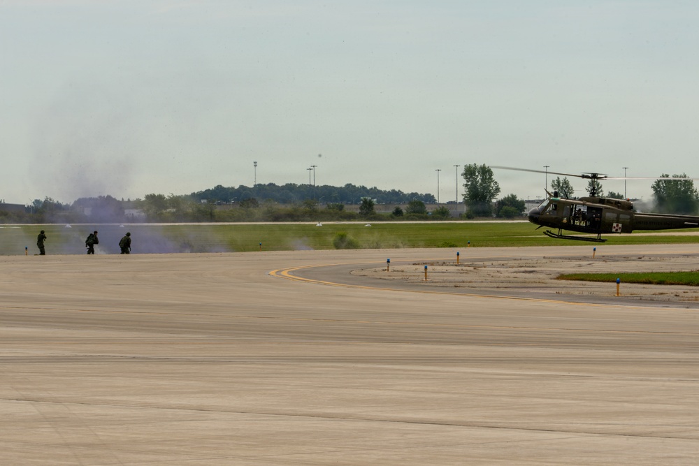 A military exercise or demonstration is taking place on an airfield tarmac. Several soldiers in combat gear are moving in a crouched formation amid smoke, while an olive-drab military helicopter (consistent with a UH-1 'Huey' type) is positioned on the right side of the tarmac. Colored smoke grenades appear to have been deployed, creating purple and white smoke clouds near the personnel.