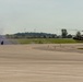 A military exercise or demonstration is taking place on an airfield tarmac. Several soldiers in combat gear are moving in a crouched formation amid smoke, while an olive-drab military helicopter (consistent with a UH-1 'Huey' type) is positioned on the right side of the tarmac. Colored smoke grenades appear to have been deployed, creating purple and white smoke clouds near the personnel.