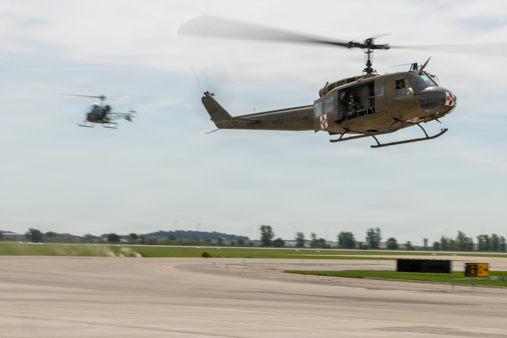 Two United States Army UH-1 'Huey' helicopters are captured in low-altitude flight over what appears to be a military airfield or airport tarmac. The foreground helicopter is clearly marked 'UNITED STATES ARMY' and displays a red cross medical insignia, indicating a medevac configuration. A second helicopter is visible in the background at a greater distance.