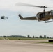 Two United States Army UH-1 'Huey' helicopters are captured in low-altitude flight over what appears to be a military airfield or airport tarmac. The foreground helicopter is clearly marked 'UNITED STATES ARMY' and displays a red cross medical insignia, indicating a medevac configuration. A second helicopter is visible in the background at a greater distance.