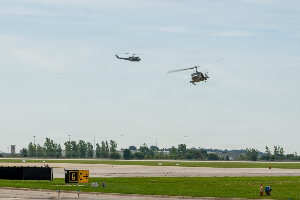 Two military helicopters, appearing to be UH-1 'Huey' type aircraft in olive drab/military green livery, are flying low over an airport or airfield. The scene appears to be a military demonstration or training exercise at a civilian or joint-use airfield. Ground infrastructure including taxiway signage and runway markings are visible in the foreground.
