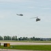 Two military helicopters, appearing to be UH-1 'Huey' type aircraft in olive drab/military green livery, are flying low over an airport or airfield. The scene appears to be a military demonstration or training exercise at a civilian or joint-use airfield. Ground infrastructure including taxiway signage and runway markings are visible in the foreground.