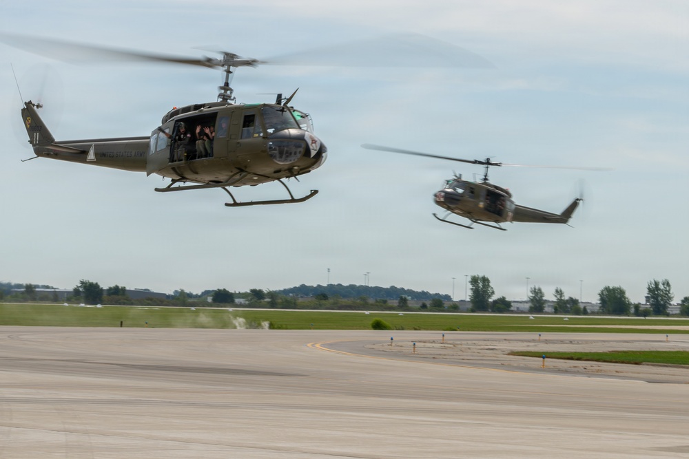 Two olive drab military helicopters, identifiable as Bell UH-1 'Huey' variants marked 'United States Army,' are flying low over an airport tarmac. The foreground helicopter has its side door open with occupants visible, one appearing to wave. A second helicopter follows in the background, slightly out of focus.