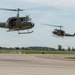 Two olive drab military helicopters, identifiable as Bell UH-1 'Huey' variants marked 'United States Army,' are flying low over an airport tarmac. The foreground helicopter has its side door open with occupants visible, one appearing to wave. A second helicopter follows in the background, slightly out of focus.