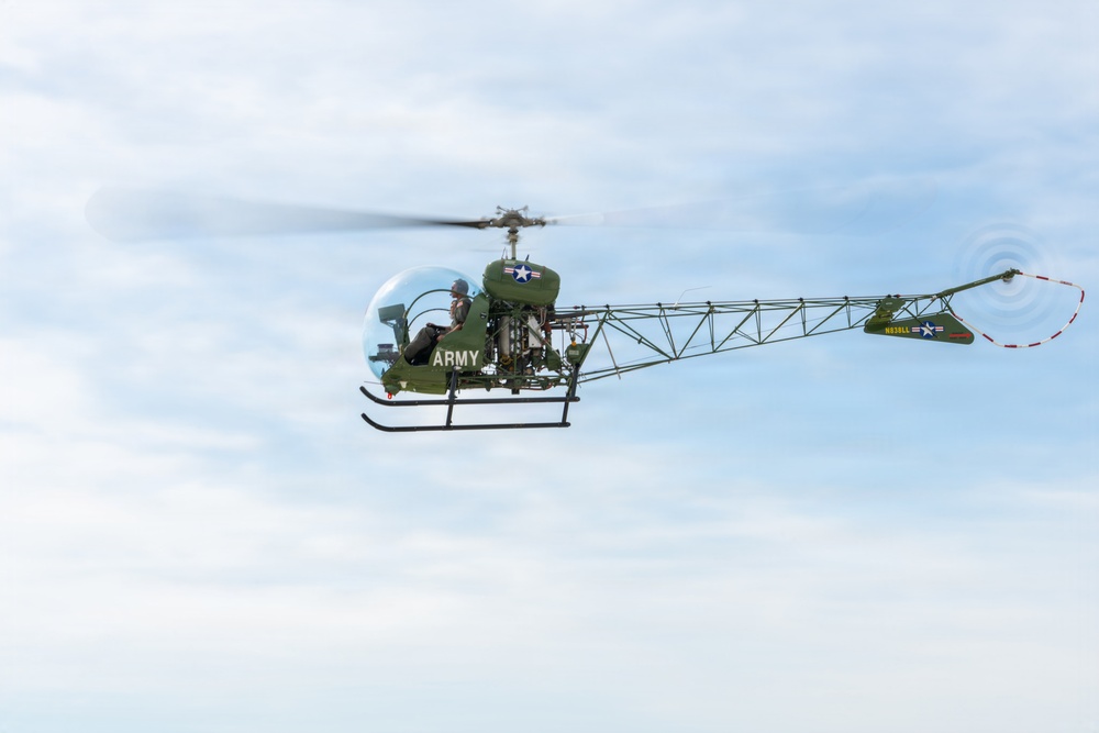 A vintage U.S. Army helicopter, identifiable as a Bell H-13 Sioux (or similar bubble-canopy light observation helicopter), is captured in flight against a partly cloudy sky. The aircraft is painted in olive drab green with U.S. military markings including a star insignia and the word 'ARMY' on the fuselage. The open lattice tail boom and bubble cockpit are characteristic features of this classic Korean War-era military helicopter.