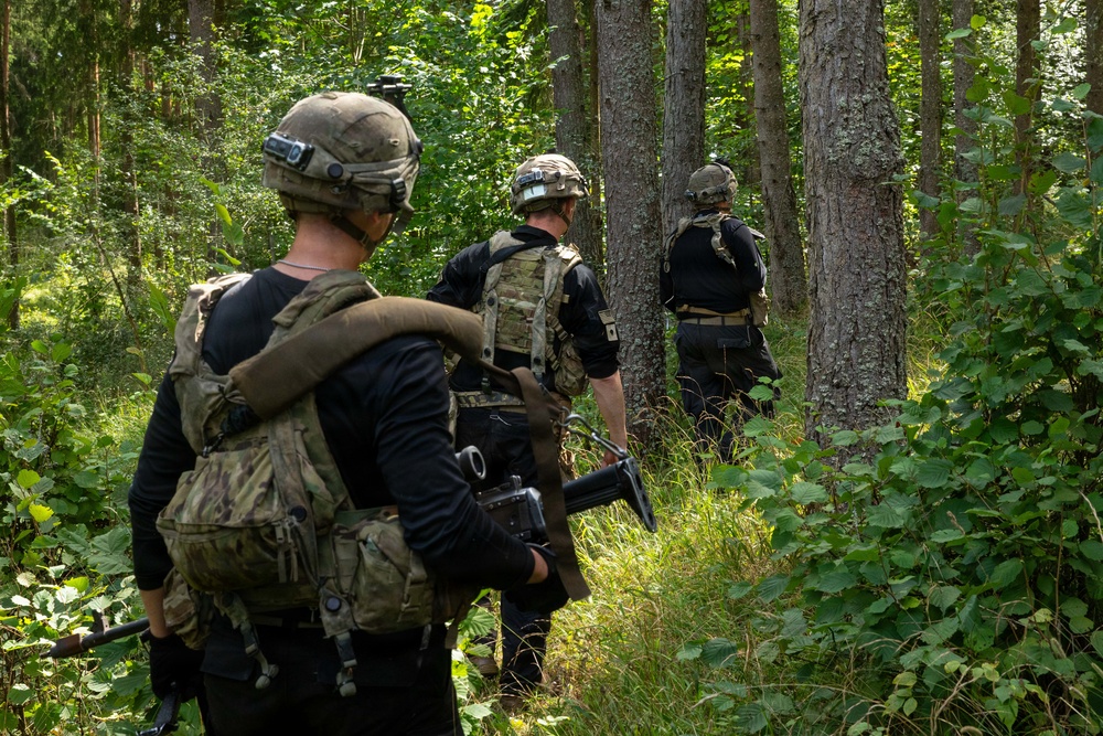 Three armed individuals in tactical military gear are moving through a dense forest in single-file formation. All three wear helmets, plate carriers or tactical vests, and black long-sleeve shirts, carrying firearms. The setting appears to be a daylight woodland patrol or training exercise.