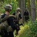 Three armed individuals in tactical military gear are moving through a dense forest in single-file formation. All three wear helmets, plate carriers or tactical vests, and black long-sleeve shirts, carrying firearms. The setting appears to be a daylight woodland patrol or training exercise.