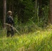 A single armed soldier in full tactical gear stands at the edge of a dense forest, appearing to observe or scan the surrounding area. The soldier is equipped with a helmet, body armor, and carries a rifle. The setting is a lush, green woodland environment with tall grass in the foreground.