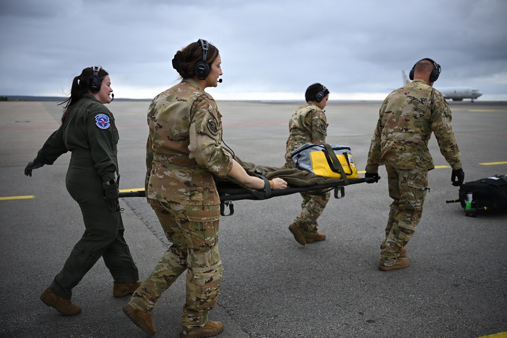 Four military personnel are carrying an empty litter/stretcher across an airfield tarmac. Three are wearing OCP (Operational Camouflage Pattern) uniforms while one wears an olive green flight suit with a squadron patch. All personnel are wearing aviation headsets, suggesting active communication during what appears to be a medical evacuation drill or operation.