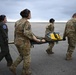 Four military personnel are carrying an empty litter/stretcher across an airfield tarmac. Three are wearing OCP (Operational Camouflage Pattern) uniforms while one wears an olive green flight suit with a squadron patch. All personnel are wearing aviation headsets, suggesting active communication during what appears to be a medical evacuation drill or operation.