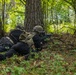 Two armed personnel in tactical gear are lying prone behind a large tree in a forested environment, with one individual aiming a rifle forward. Both are wearing black clothing, tactical vests, helmets, and backpacks, suggesting a military training exercise or operational scenario. The setting is a deciduous forest with dense green foliage and dappled natural light.