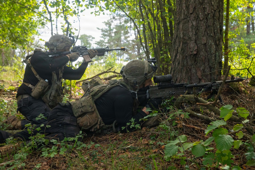 Two armed personnel in tactical gear are positioned in a wooded area, taking up defensive or offensive firing positions behind a large tree. One individual is prone on the ground aiming a long-barreled rifle with a scope, while the second is in a kneeling position aiming a shorter carbine-style rifle. Both wear multicam/camouflage plate carriers, helmets with mounted accessories, and black base layers.