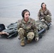 Two U.S. Air Force personnel in OCP (Operational Camouflage Pattern) uniforms are kneeling on a tarmac or airfield surface, each equipped with communication headsets. They are positioned beside military litters (stretchers) that appear to contain simulated casualties, suggesting an aeromedical evacuation training exercise. The foreground airman's name tape reads 'JONES' and the uniform patch reads 'U.S. AIR FORCE'.