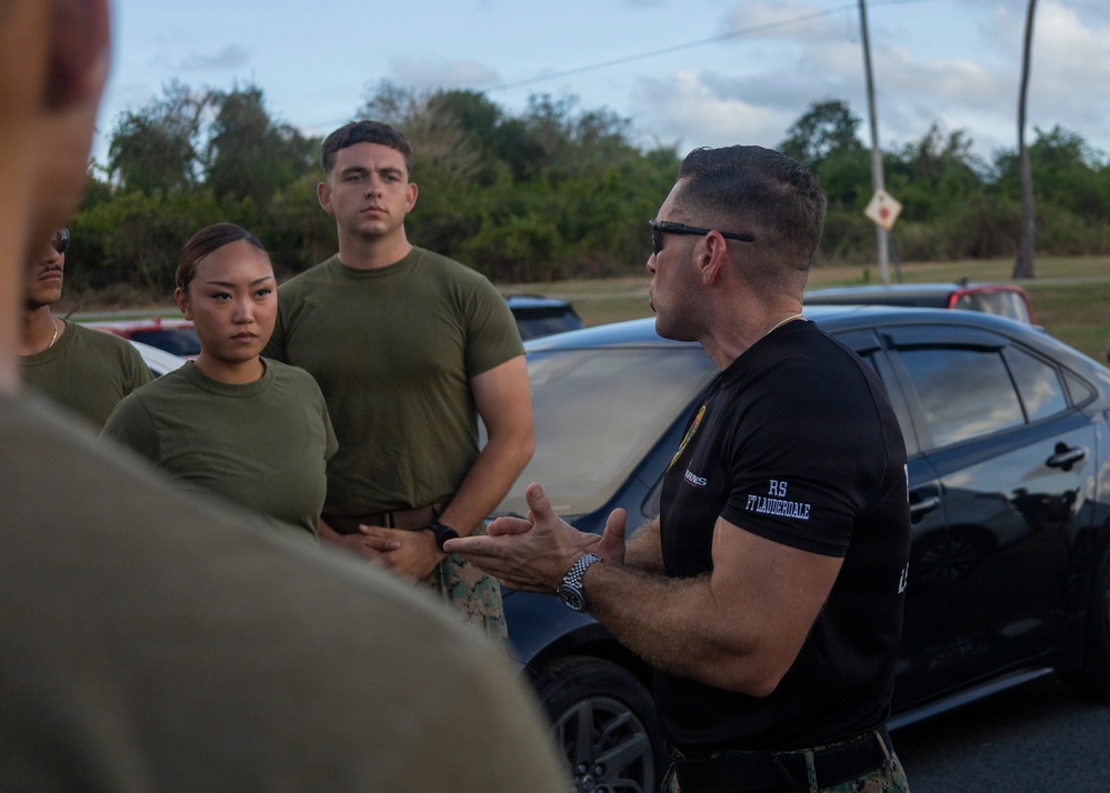 A man in a black t-shirt with 'RS FT LAUDERDALE' text is speaking and gesturing with his hands to a group of individuals wearing olive green military-style t-shirts. The setting appears to be an outdoor parking area or roadside location during daylight hours. The scene suggests a briefing or instructional session.