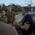 LCF-24 and RS Fort Lauderdale Poolee Function A man in a black t-shirt with 'RS FT LAUDERDALE' text is speaking and gesturing with his hands to a group of individuals wearing olive green military-style t-shirts. The setting appears to be an outdoor parking area or roadside location during daylight hours. The scene suggests a briefing or instructional session.