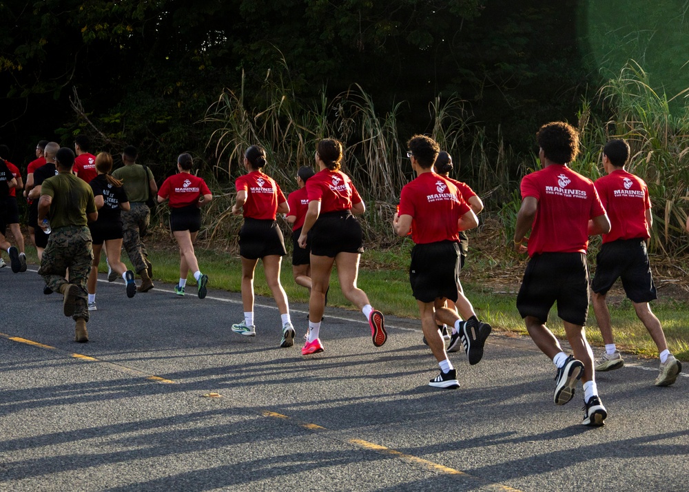 A group of individuals wearing red 'Marines: The Few The Proud' t-shirts and black shorts are running on a paved road alongside personnel in military camouflage uniforms. The scene appears to be a physical training or recruiting event, likely a Marine Corps fitness run. The image is taken from behind the runners, with dense green vegetation visible in the background.