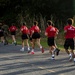 A group of individuals wearing red 'Marines: The Few The Proud' t-shirts and black shorts are running on a paved road alongside personnel in military camouflage uniforms. The scene appears to be a physical training or recruiting event, likely a Marine Corps fitness run. The image is taken from behind the runners, with dense green vegetation visible in the background.