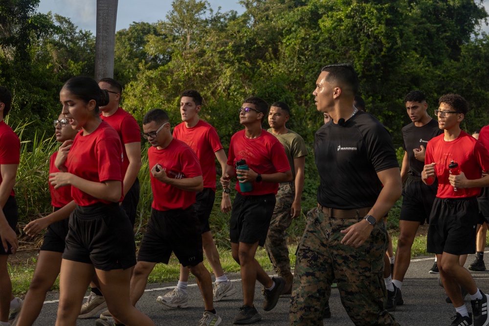 A group of young individuals in red t-shirts and black shorts are participating in a physical training run or march on a paved road, led by a U.S. Marine in a black 'Marines' t-shirt and camouflage utility trousers. The setting appears to be a military base or training facility surrounded by dense tropical vegetation. The scene suggests a recruiting or JROTC-style physical fitness event.