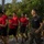 LCF-24 and RS Fort Lauderdale Poolee Function A group of young individuals in red t-shirts and black shorts are participating in a physical training run or march on a paved road, led by a U.S. Marine in a black 'Marines' t-shirt and camouflage utility trousers. The setting appears to be a military base or training facility surrounded by dense tropical vegetation. The scene suggests a recruiting or JROTC-style physical fitness event.