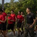 A group of young individuals in red t-shirts and black shorts are participating in a physical training run or march on a paved road, led by a U.S. Marine in a black 'Marines' t-shirt and camouflage utility trousers. The setting appears to be a military base or training facility surrounded by dense tropical vegetation. The scene suggests a recruiting or JROTC-style physical fitness event.
