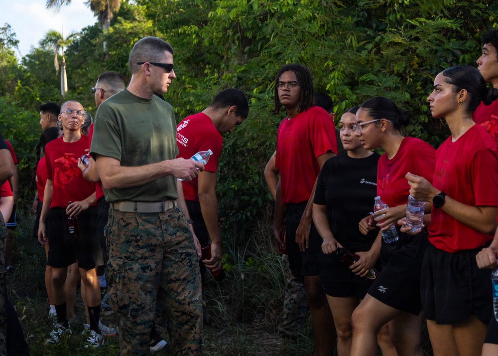A U.S. Marine in olive drab t-shirt and camouflage utilities is addressing a group of young recruits or poolees wearing red Marines t-shirts and black athletic shorts. The setting appears to be an outdoor area with dense tropical vegetation, suggesting a training or orientation event. Several participants are holding water bottles, indicating a post-physical activity rest or briefing period.