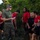 LCF-24 and RS Fort Lauderdale Poolee Function A U.S. Marine in olive drab t-shirt and camouflage utilities is addressing a group of young recruits or poolees wearing red Marines t-shirts and black athletic shorts. The setting appears to be an outdoor area with dense tropical vegetation, suggesting a training or orientation event. Several participants are holding water bottles, indicating a post-physical activity rest or briefing period.