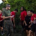 A U.S. Marine in olive drab t-shirt and camouflage utilities is addressing a group of young recruits or poolees wearing red Marines t-shirts and black athletic shorts. The setting appears to be an outdoor area with dense tropical vegetation, suggesting a training or orientation event. Several participants are holding water bottles, indicating a post-physical activity rest or briefing period.