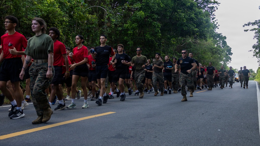 A large group of military personnel and civilians are running together along a paved road lined with dense green trees. The group includes individuals in military camouflage uniforms mixed with civilians wearing red and black t-shirts, suggesting a joint military-community fitness run or recruitment event. A vehicle follows the group from behind, likely providing safety escort.
