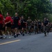 A large group of military personnel and civilians are running together along a paved road lined with dense green trees. The group includes individuals in military camouflage uniforms mixed with civilians wearing red and black t-shirts, suggesting a joint military-community fitness run or recruitment event. A vehicle follows the group from behind, likely providing safety escort.