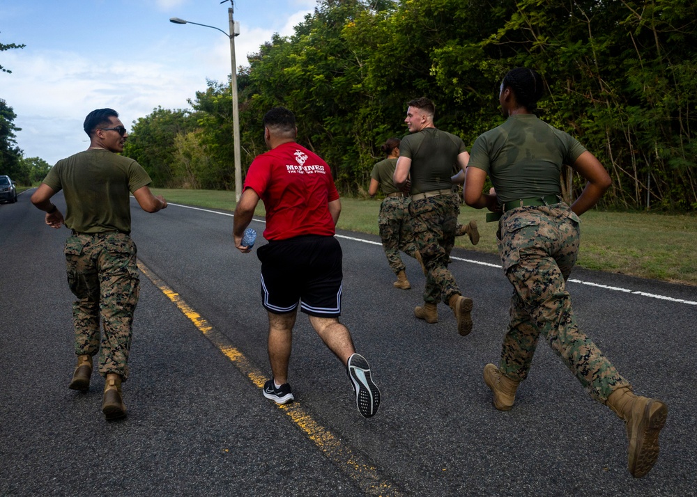 A group of U.S. Marines in military camouflage uniforms and olive drab t-shirts are running together along a paved road bordered by dense tropical vegetation. One individual stands out wearing a red 'Marines' branded t-shirt and black shorts, suggesting a mix of uniformed and civilian/PT attire. The setting appears to be a military base or installation in a tropical environment, likely during a physical training exercise.