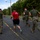 LCF-24 and RS Fort Lauderdale Poolee Function A group of U.S. Marines in military camouflage uniforms and olive drab t-shirts are running together along a paved road bordered by dense tropical vegetation. One individual stands out wearing a red 'Marines' branded t-shirt and black shorts, suggesting a mix of uniformed and civilian/PT attire. The setting appears to be a military base or installation in a tropical environment, likely during a physical training exercise.