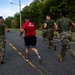 A group of U.S. Marines in military camouflage uniforms and olive drab t-shirts are running together along a paved road bordered by dense tropical vegetation. One individual stands out wearing a red 'Marines' branded t-shirt and black shorts, suggesting a mix of uniformed and civilian/PT attire. The setting appears to be a military base or installation in a tropical environment, likely during a physical training exercise.