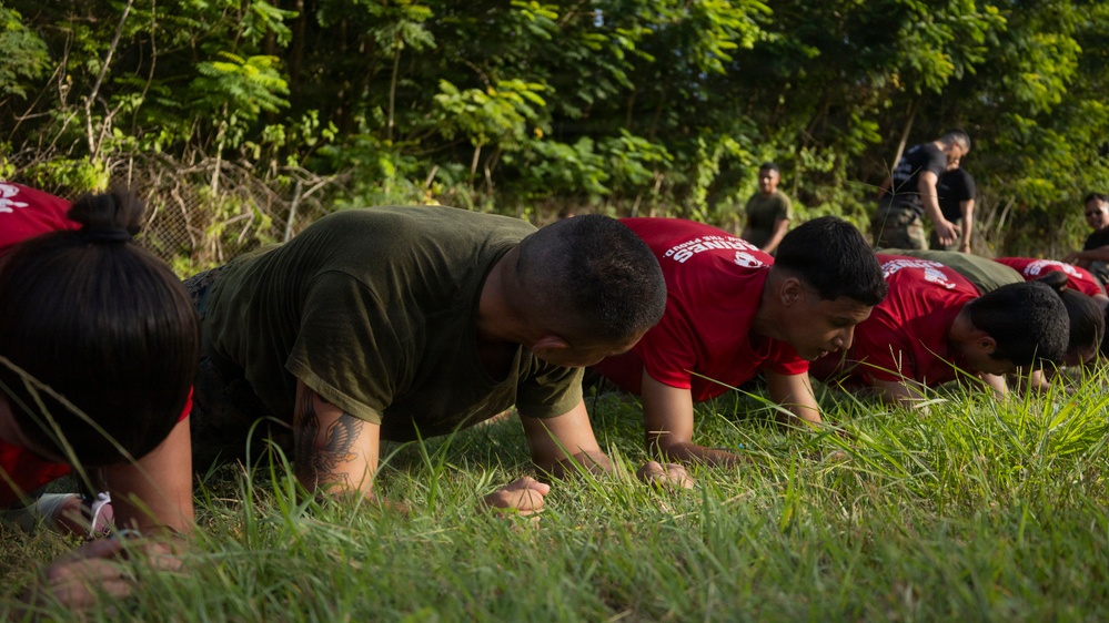 A group of individuals in red and olive/green t-shirts are performing push-ups or a low plank exercise on a grassy outdoor area. The setting appears to be a military or fitness training event, with supervisors or instructors visible standing in the background. The participants are aligned in a row, suggesting a structured group physical training session.