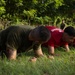 A group of individuals in red and olive/green t-shirts are performing push-ups or a low plank exercise on a grassy outdoor area. The setting appears to be a military or fitness training event, with supervisors or instructors visible standing in the background. The participants are aligned in a row, suggesting a structured group physical training session.