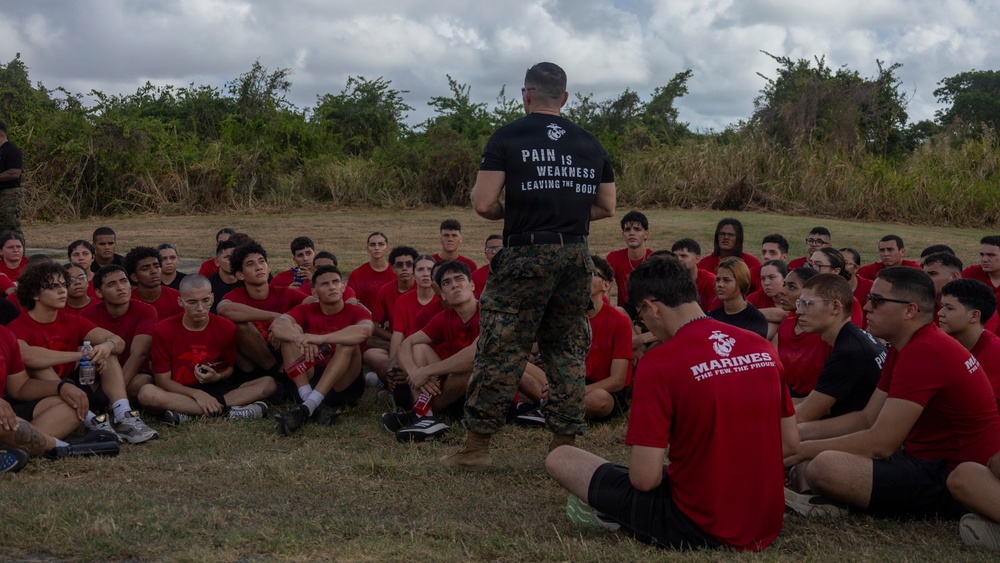 A U.S. Marine instructor in camouflage trousers and a black 'Pain is Weakness Leaving the Body' t-shirt addresses a large group of young recruits or poolees seated on grass in a semicircle. The recruits are wearing matching red t-shirts, several bearing the Marines 'The Few The Proud' logo. The setting appears to be an outdoor training or orientation event, likely a recruiting function or poolee function.