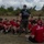 LCF-24 and RS Fort Lauderdale Poolee Function A U.S. Marine instructor in camouflage trousers and a black 'Pain is Weakness Leaving the Body' t-shirt addresses a large group of young recruits or poolees seated on grass in a semicircle. The recruits are wearing matching red t-shirts, several bearing the Marines 'The Few The Proud' logo. The setting appears to be an outdoor training or orientation event, likely a recruiting function or poolee function.