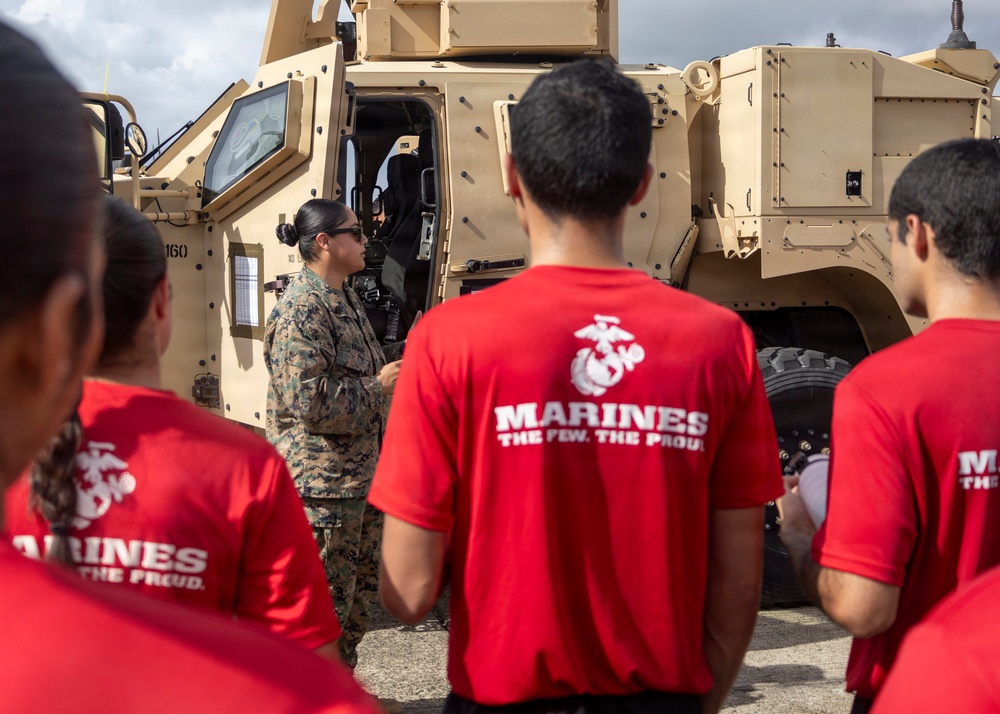 A U.S. Marine in camouflage uniform is briefing a group of civilians wearing red 'Marines - The Few, The Proud' t-shirts in front of a tan MRAP (Mine-Resistant Ambush Protected) military vehicle. The setting appears to be an outdoor military demonstration or recruiting event. The group is attentively listening to the Marine's presentation near the open door of the armored vehicle.