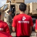 A U.S. Marine in camouflage uniform is briefing a group of civilians wearing red 'Marines - The Few, The Proud' t-shirts in front of a tan MRAP (Mine-Resistant Ambush Protected) military vehicle. The setting appears to be an outdoor military demonstration or recruiting event. The group is attentively listening to the Marine's presentation near the open door of the armored vehicle.