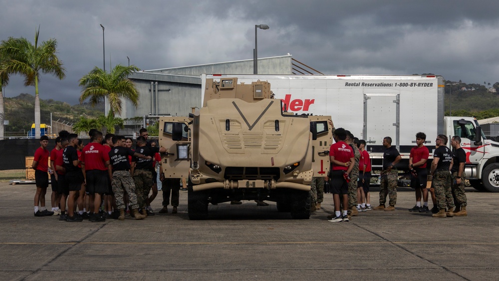 A group of individuals in red and black shirts, along with personnel in military camouflage uniforms, are gathered around a tan/desert-colored military armored vehicle (appears to be a JLTV or MRAP-type vehicle) on a paved lot. The setting appears to be a port or industrial area in a tropical location, with palm trees and hills visible in the background. A Ryder rental truck is parked behind the military vehicle.