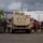 LCF-24 and RS Fort Lauderdale Poolee Function A group of individuals in red and black shirts, along with personnel in military camouflage uniforms, are gathered around a tan/desert-colored military armored vehicle (appears to be a JLTV or MRAP-type vehicle) on a paved lot. The setting appears to be a port or industrial area in a tropical location, with palm trees and hills visible in the background. A Ryder rental truck is parked behind the military vehicle.