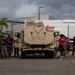 A group of individuals in red and black shirts, along with personnel in military camouflage uniforms, are gathered around a tan/desert-colored military armored vehicle (appears to be a JLTV or MRAP-type vehicle) on a paved lot. The setting appears to be a port or industrial area in a tropical location, with palm trees and hills visible in the background. A Ryder rental truck is parked behind the military vehicle.