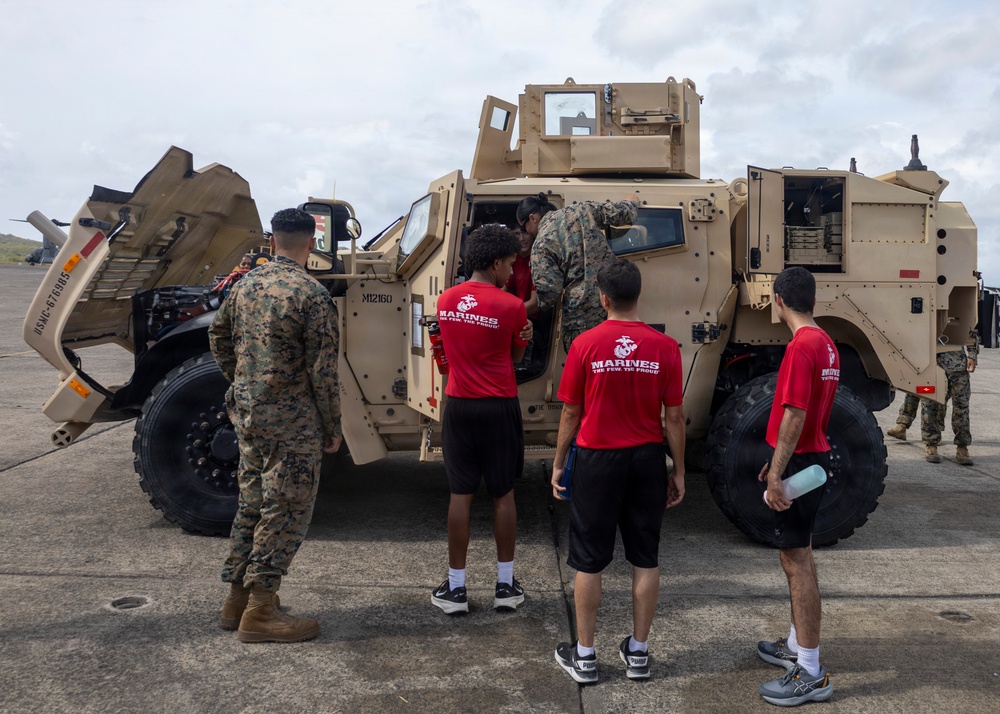 A group of civilians wearing red 'Marines: The Few, The Proud' shirts are being shown a tan/desert-colored military JLTV (Joint Light Tactical Vehicle) by a uniformed Marine in camouflage on what appears to be a tarmac or airfield. One civilian is climbing into or inspecting the vehicle's cab while others observe. This appears to be a Marine Corps recruiting or public outreach event.