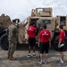 A group of civilians wearing red 'Marines: The Few, The Proud' shirts are being shown a tan/desert-colored military JLTV (Joint Light Tactical Vehicle) by a uniformed Marine in camouflage on what appears to be a tarmac or airfield. One civilian is climbing into or inspecting the vehicle's cab while others observe. This appears to be a Marine Corps recruiting or public outreach event.