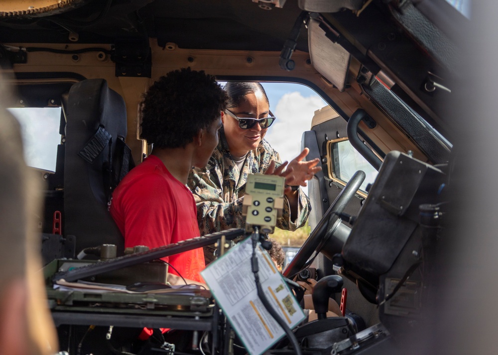 A military service member in camouflage uniform and sunglasses is explaining or demonstrating something inside a military vehicle, likely a HMMWV (Humvee), to a civilian wearing a red shirt. The service member appears to be gesturing while pointing out vehicle components or controls. This appears to be a public outreach, recruitment, or educational demonstration event.