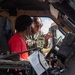 A military service member in camouflage uniform and sunglasses is explaining or demonstrating something inside a military vehicle, likely a HMMWV (Humvee), to a civilian wearing a red shirt. The service member appears to be gesturing while pointing out vehicle components or controls. This appears to be a public outreach, recruitment, or educational demonstration event.