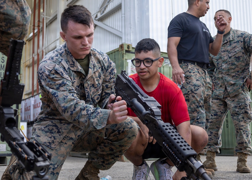 A U.S. Marine in digital camouflage uniform is demonstrating a firearm to a young civilian wearing a red shirt and glasses. The setting appears to be an outdoor military facility or base, with shipping containers and a corrugated metal building visible in the background. Additional Marines and civilians are visible in the background, suggesting a public outreach or recruitment event.